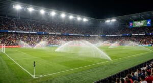 Groundskeepers watering a soccer field during halftime break, with stadium lights illuminating the empty pitch